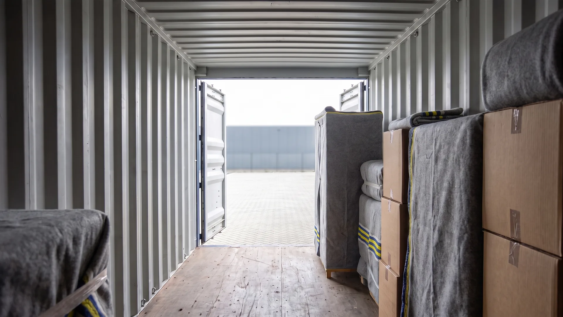 Zicht vanuit de laadruimte van een zeecontainer op de geopende deuren, met meubels in verhuisdekens en verhuisdozen en een deuropening die lager is dan het plafond.