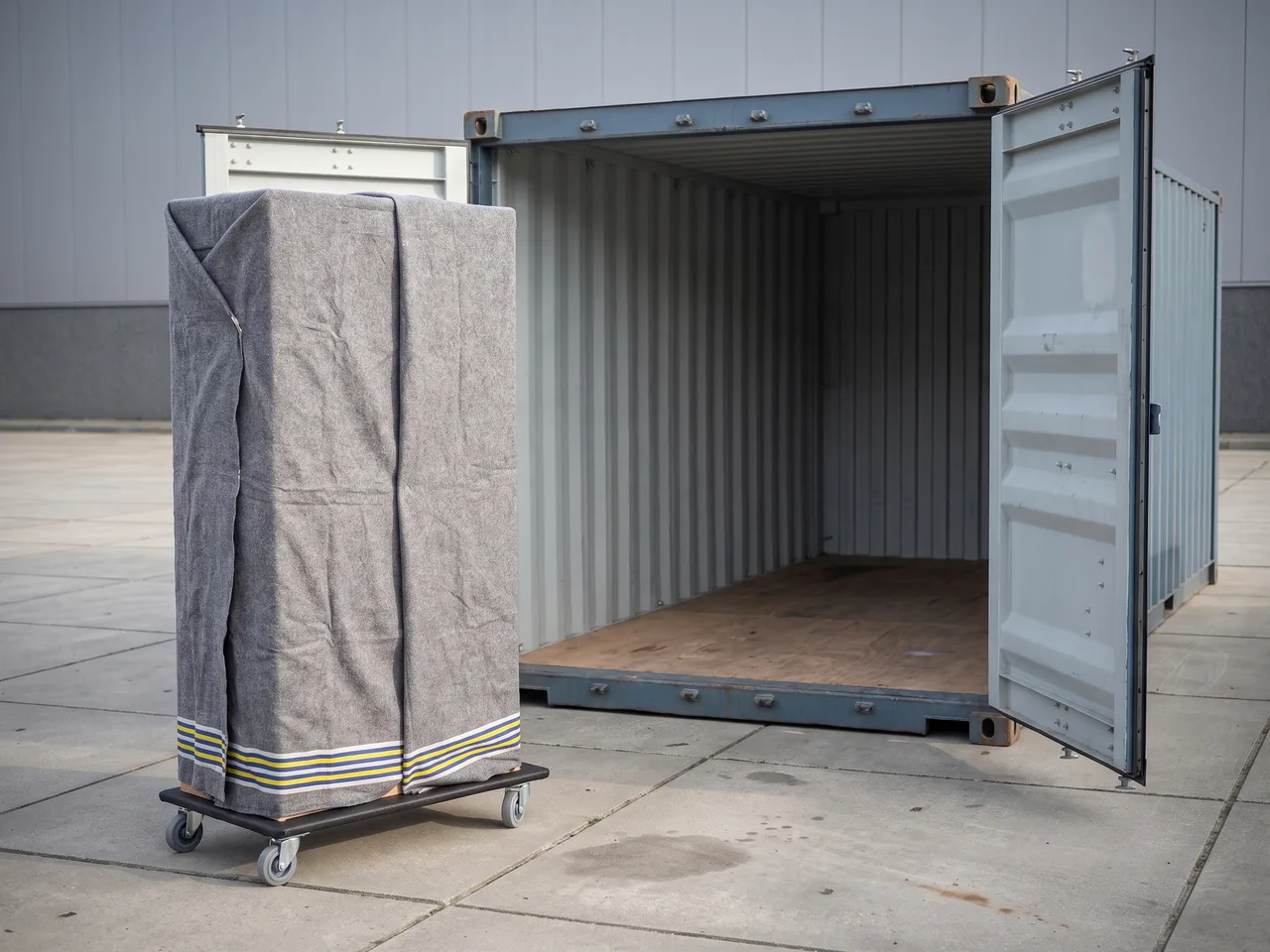 Hoge kast in verhuisdekens op een meubelhond naast de geopende deuren van een zeecontainer.