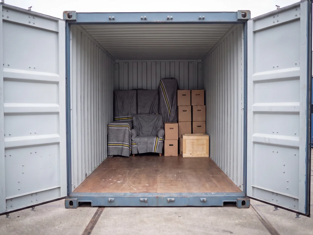 Zeecontainer met geopende deuren, achterin een kleine inboedel met ingepakte meubels, dozen en een houten krat.