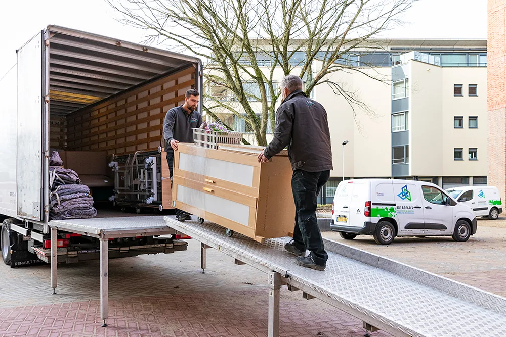 Twee verhuizers van De Bresser rijden een kast over de laadbrug uit de verhuiswagen, met twee bestelbussen van De Bresser op de achtergrond
