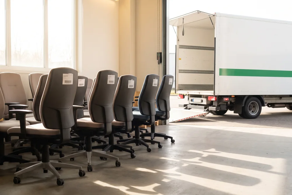 Rijen gebruikte bureaustoelen met labels in de opslaghal, met een bakwagen voor de open deur