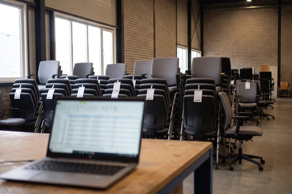 Laptop met een voorraadlijst op een werkbank, met rijen gelabelde bureaustoelen in de hal