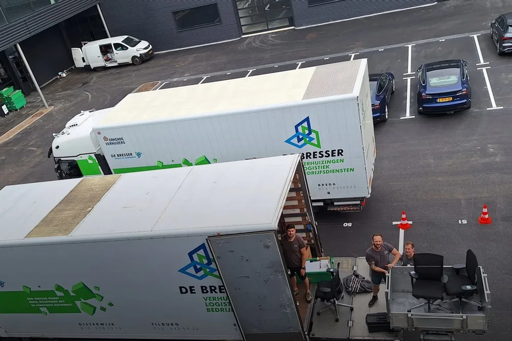 Twee vrachtwagens van De Bresser van bovenaf gezien, verhuizers zetten bureaustoelen op het plateau van de verhuislift