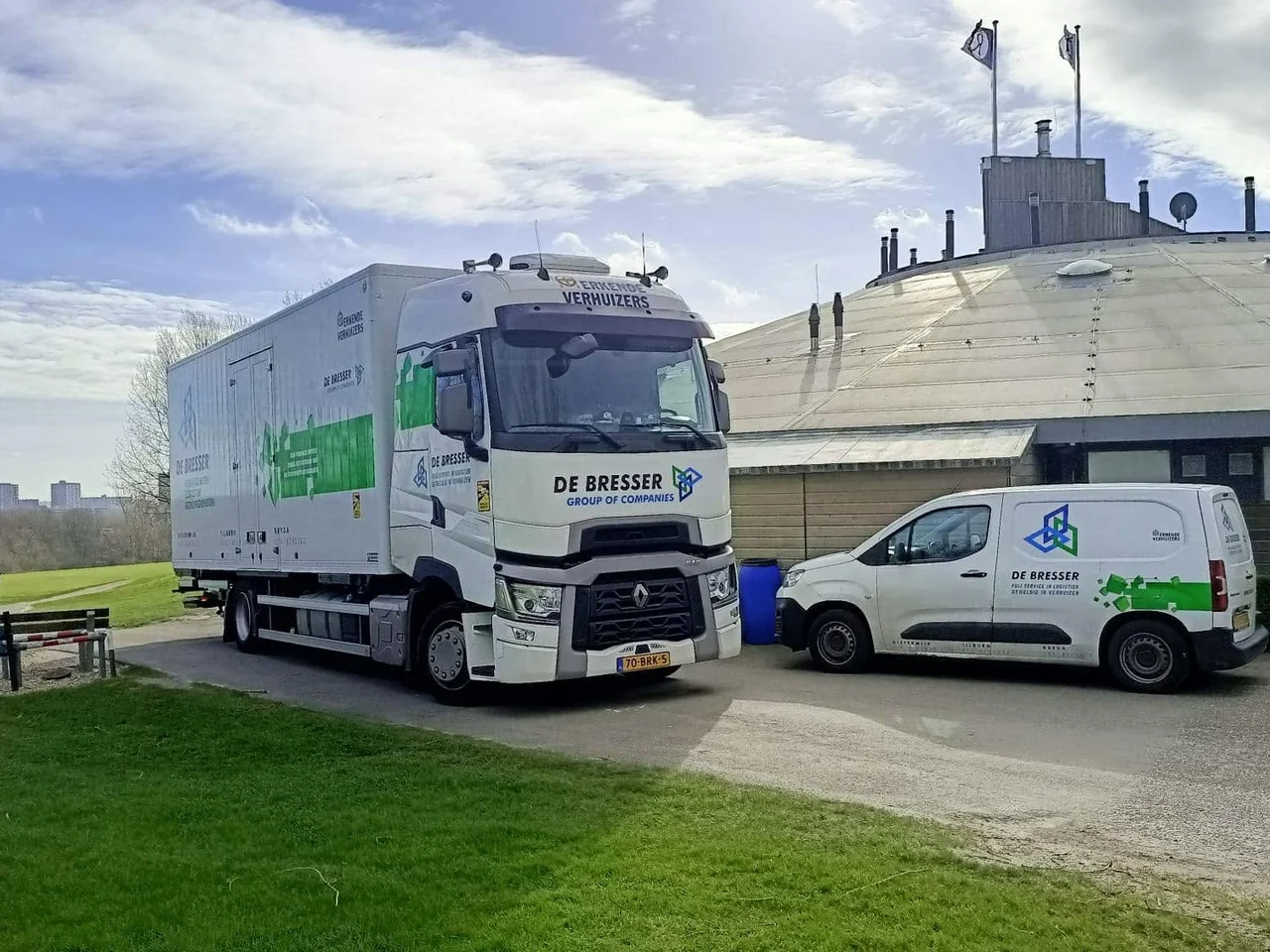 Vrachtwagen en bestelbus van De Bresser geparkeerd bij een rond gebouw aan een grasveld