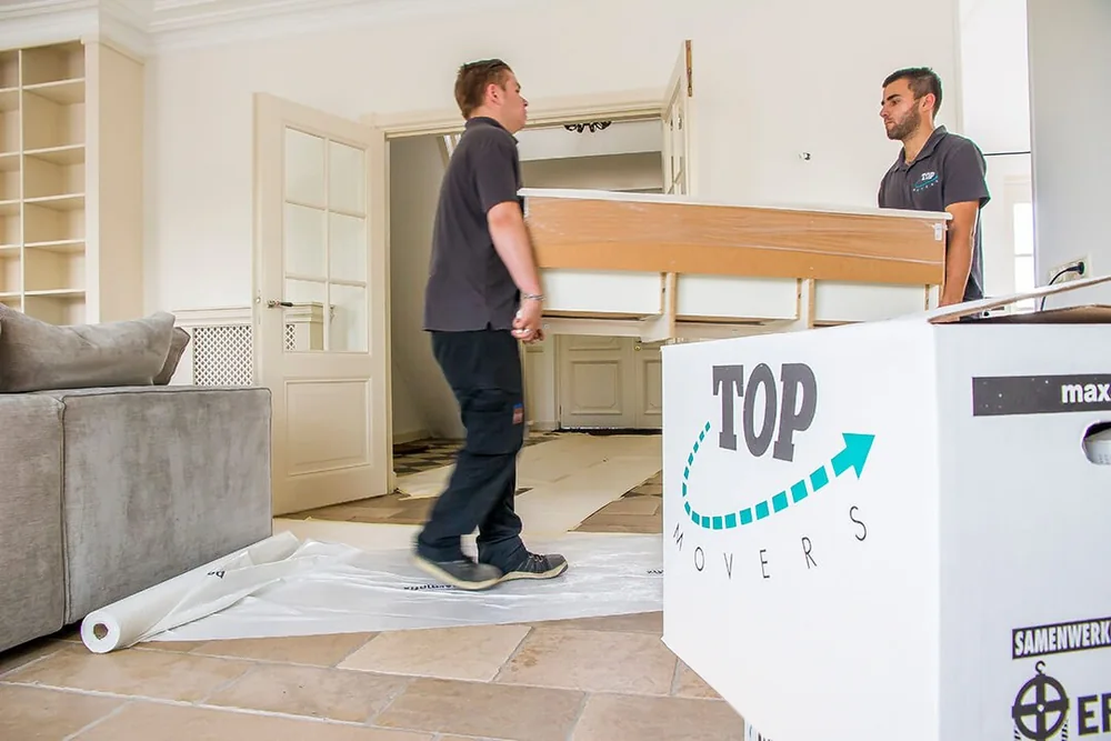 Twee verhuizers dragen een tafel door een woonkamer met afgedekte vloer en een verhuisdoos