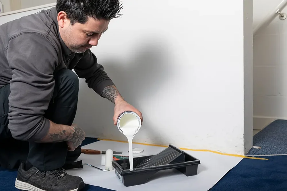 Medewerker van De Bresser giet witte verf in een verfbak op een afgeplakte vloer