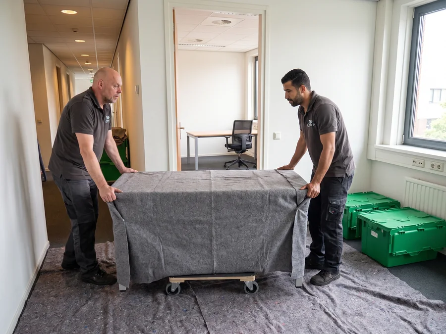 Twee verhuizers van De Bresser rijden een bureau onder een verhuisdeken op een hondje door een kantoorgang, met groene kratten ernaast