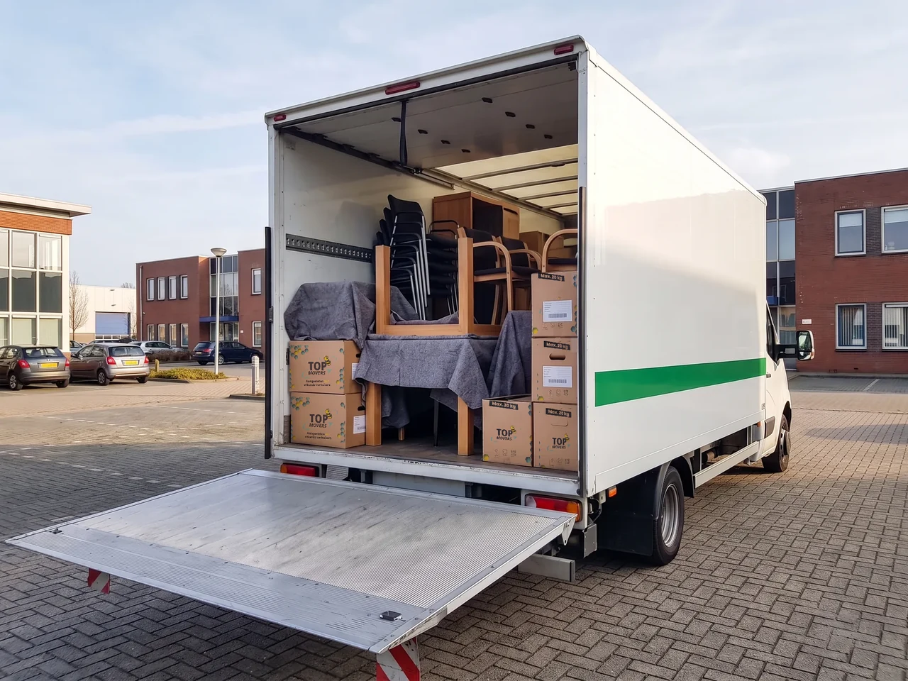 Open achterkant van een witte bakwagen met de laadklep omlaag, geladen met stoelen, in dekens verpakte tafels en dozen met labels