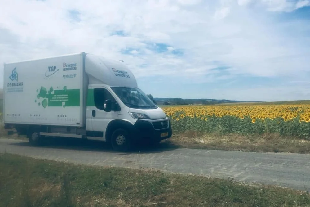 Bakwagen van De Bresser met het logo van Erkende Verhuizers op een landweg langs een veld met zonnebloemen