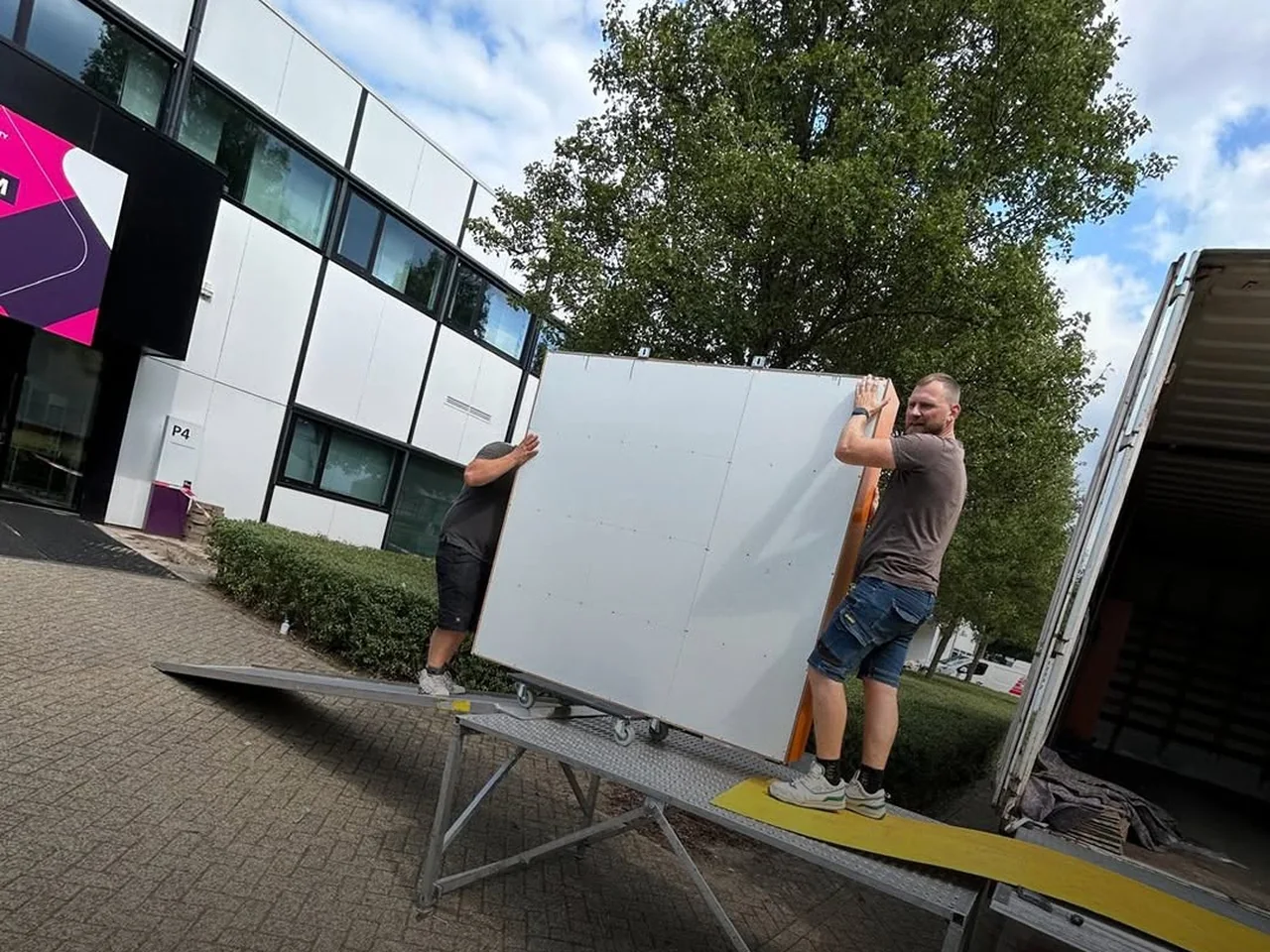 Twee medewerkers van De Bresser dragen een groot paneel over een loopplank de laadbak in
