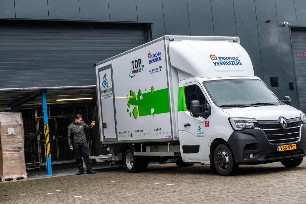 Verhuiswagen van De Bresser aan het laadperron van een bedrijfshal, met een medewerker ernaast