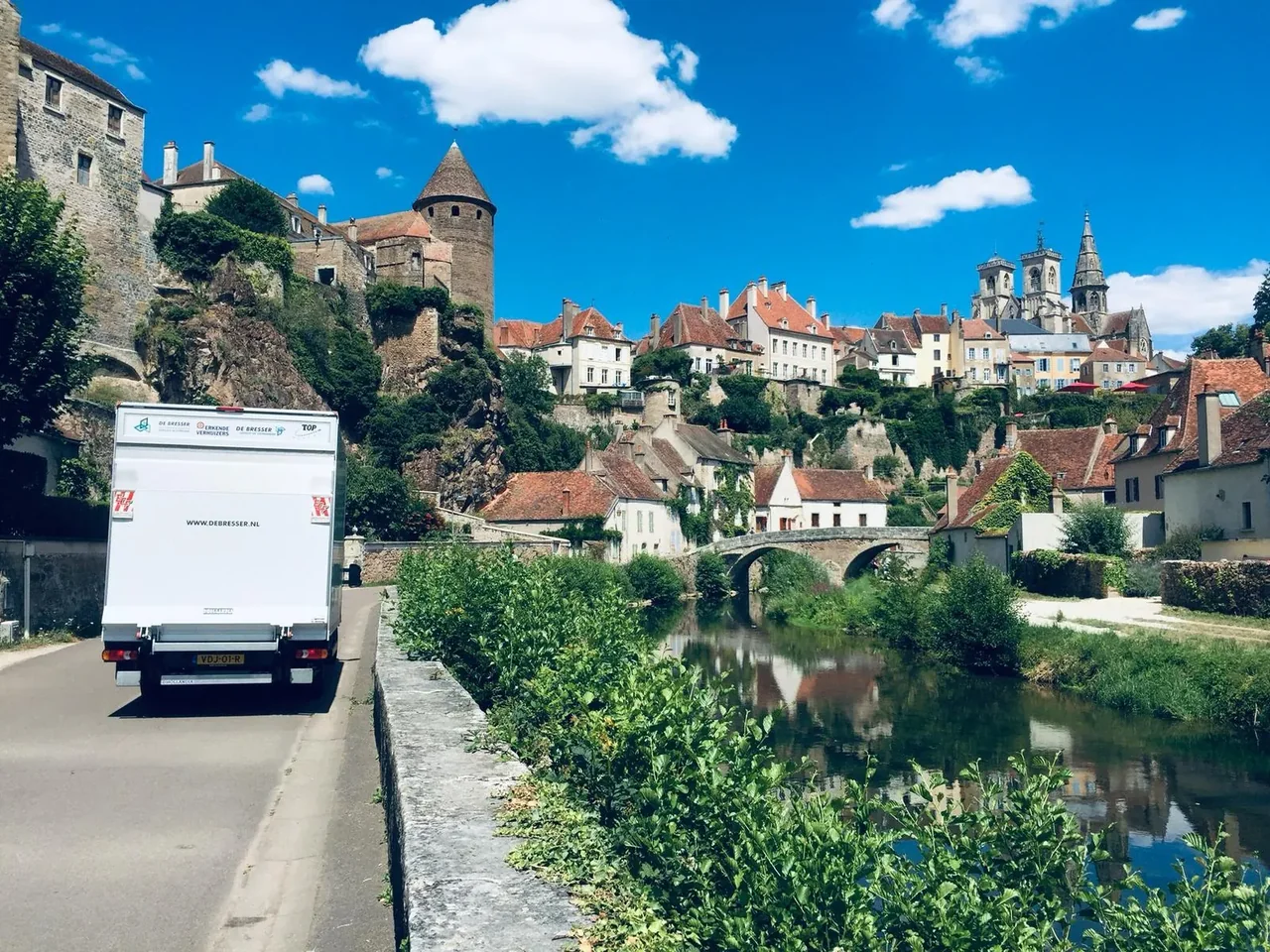 Verhuiswagen van De Bresser rijdt langs een rivier naar een oud stadje met torens en een stenen brug