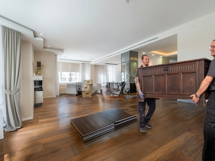 Twee verhuizers dragen een eikenhouten kist door een lichte, bijna lege woonkamer