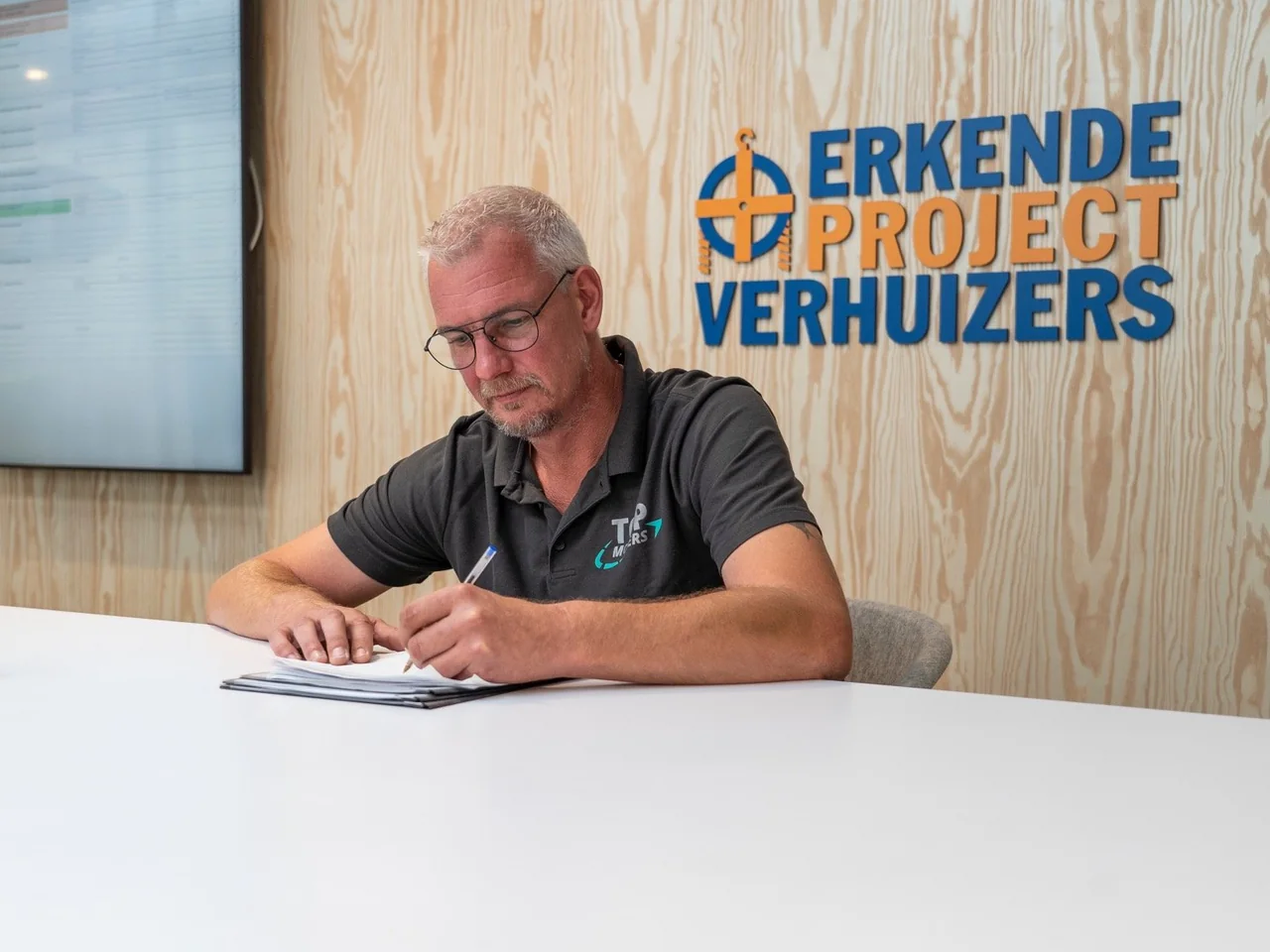 Medewerker van De Bresser aan tafel met een notitieblok, met het woordmerk Erkende Projectverhuizers op de wand