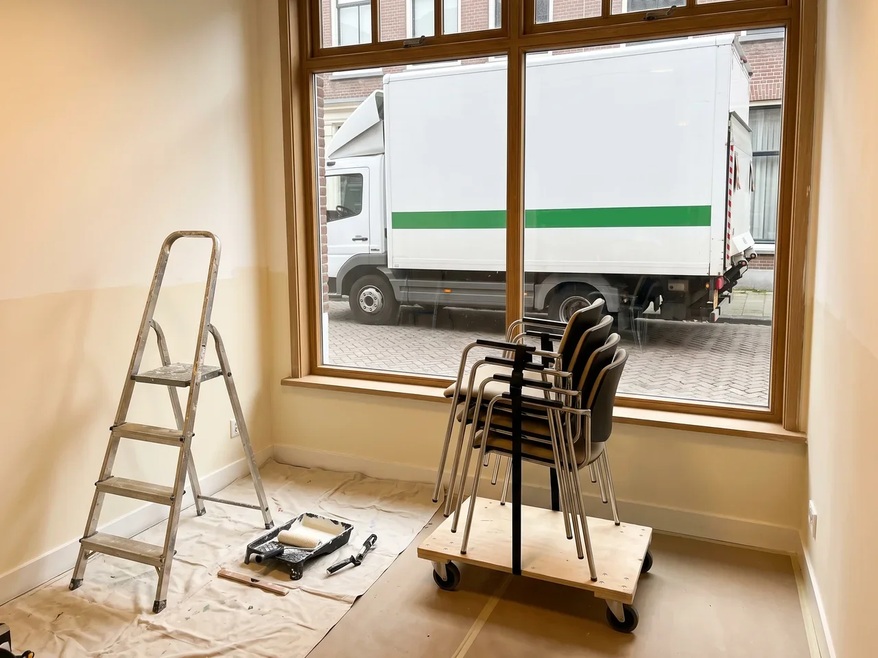 Lege kamer met een trapje, een verfbak met roller en gestapelde stoelen op een rolplateau voor een groot raam
