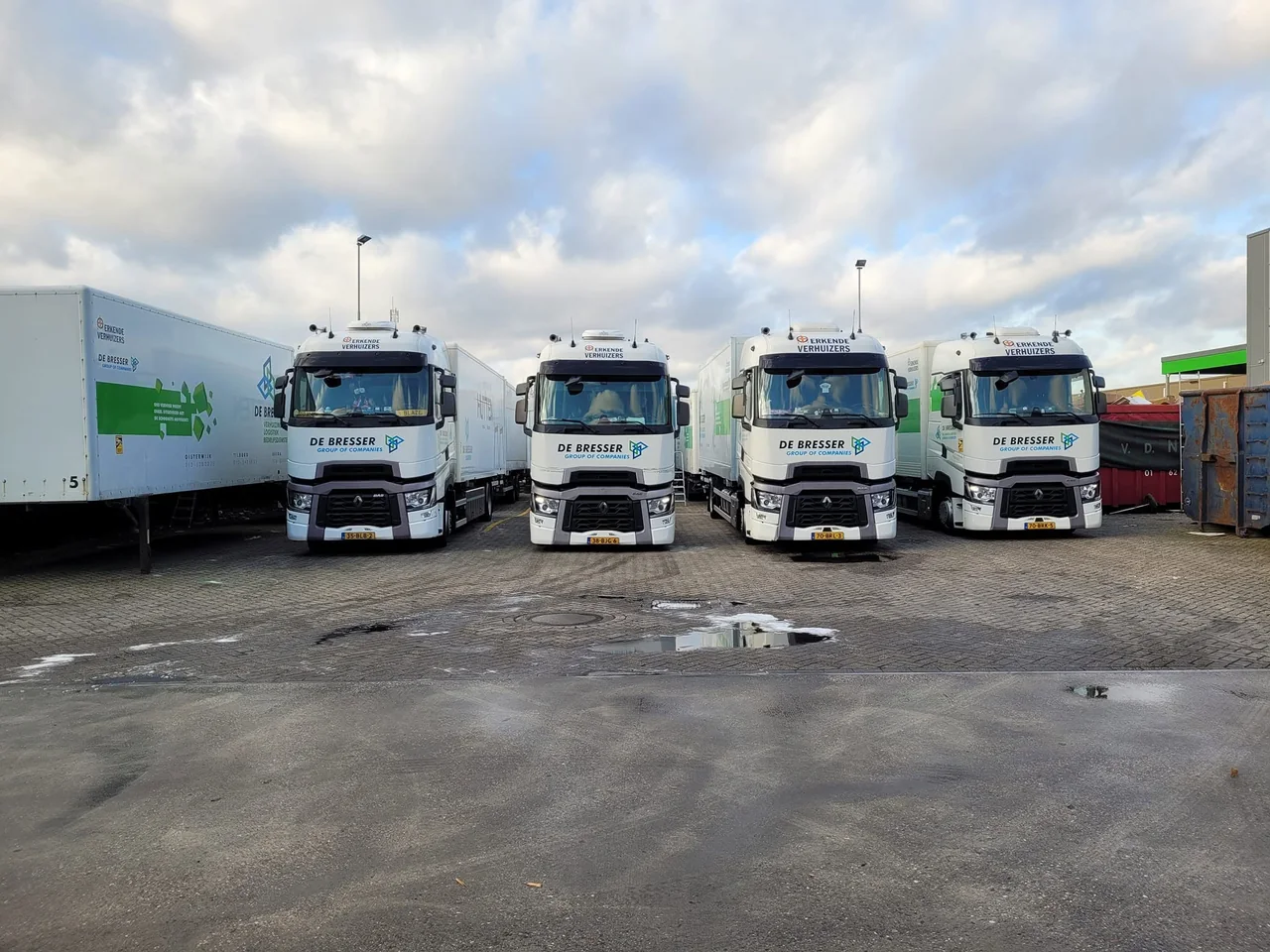 Vier witte vrachtwagens van De Bresser naast elkaar op het terrein, met een oplegger links en containers rechts
