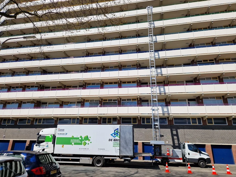 Verhuiswagen van De Bresser en een verhuislift aan een hoge galerijflat, de stoep afgezet met pionnen