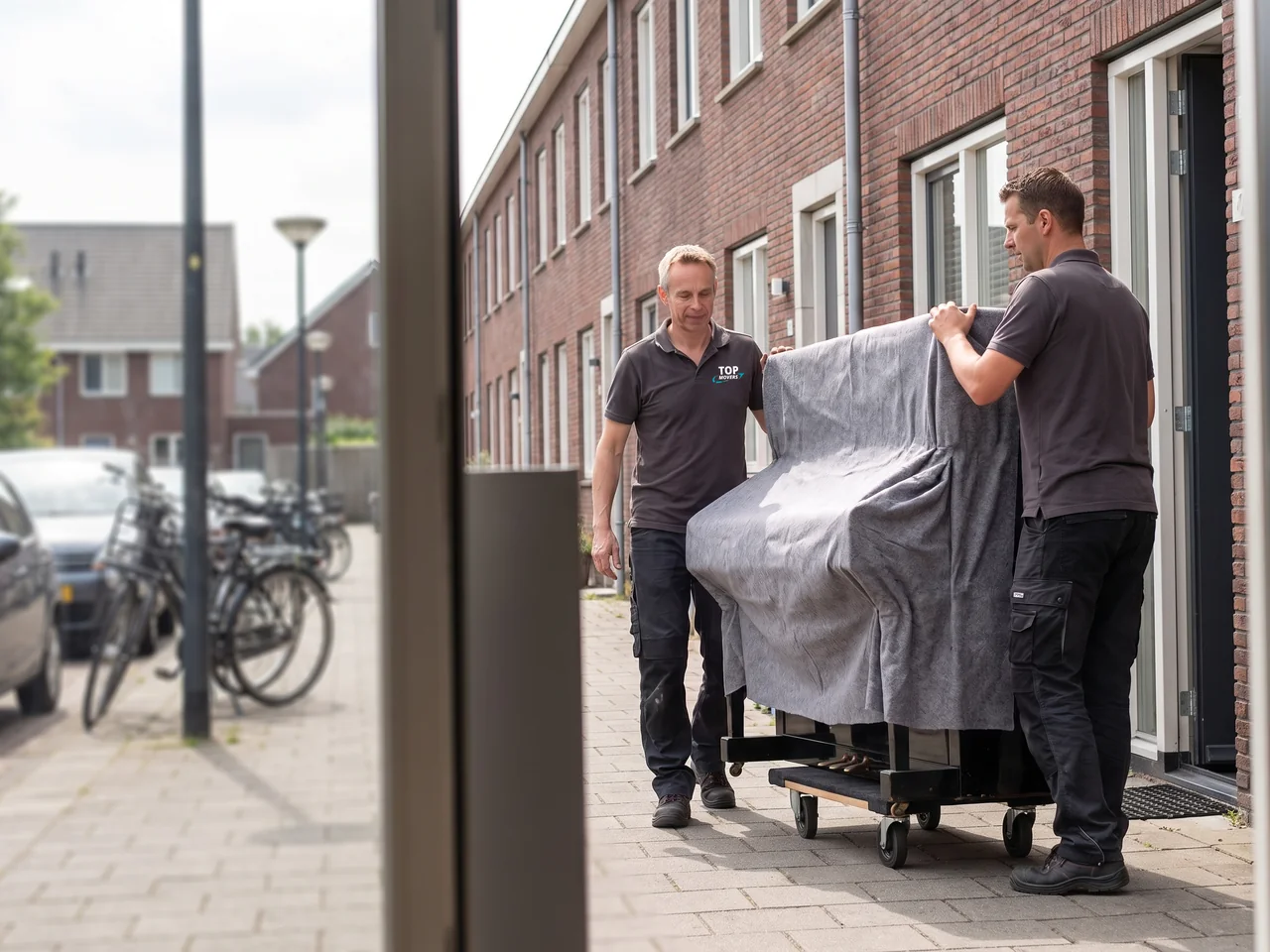 Twee verhuizers rijden een ingepakte piano op een pianokar over de stoep naar buiten