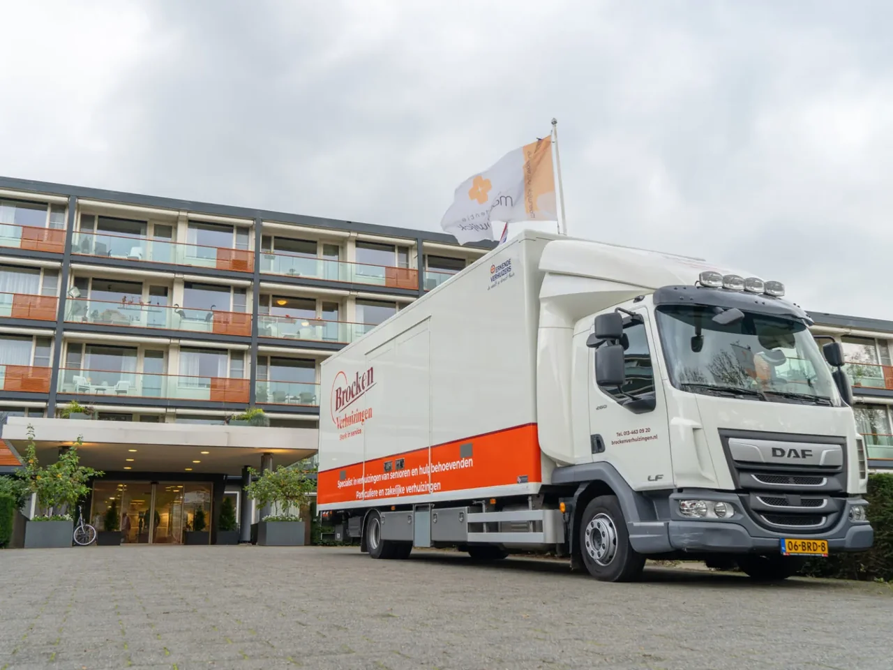 Verhuiswagen van Brocken Verhuizingen voor de ingang van een woonzorgcomplex met een vlag op het dak